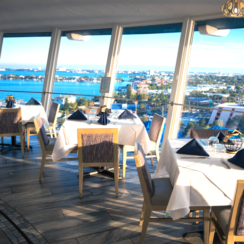 The image shows a bright restaurant with tables set by large windows, offering a scenic view of a coastal city and water body in the distance.